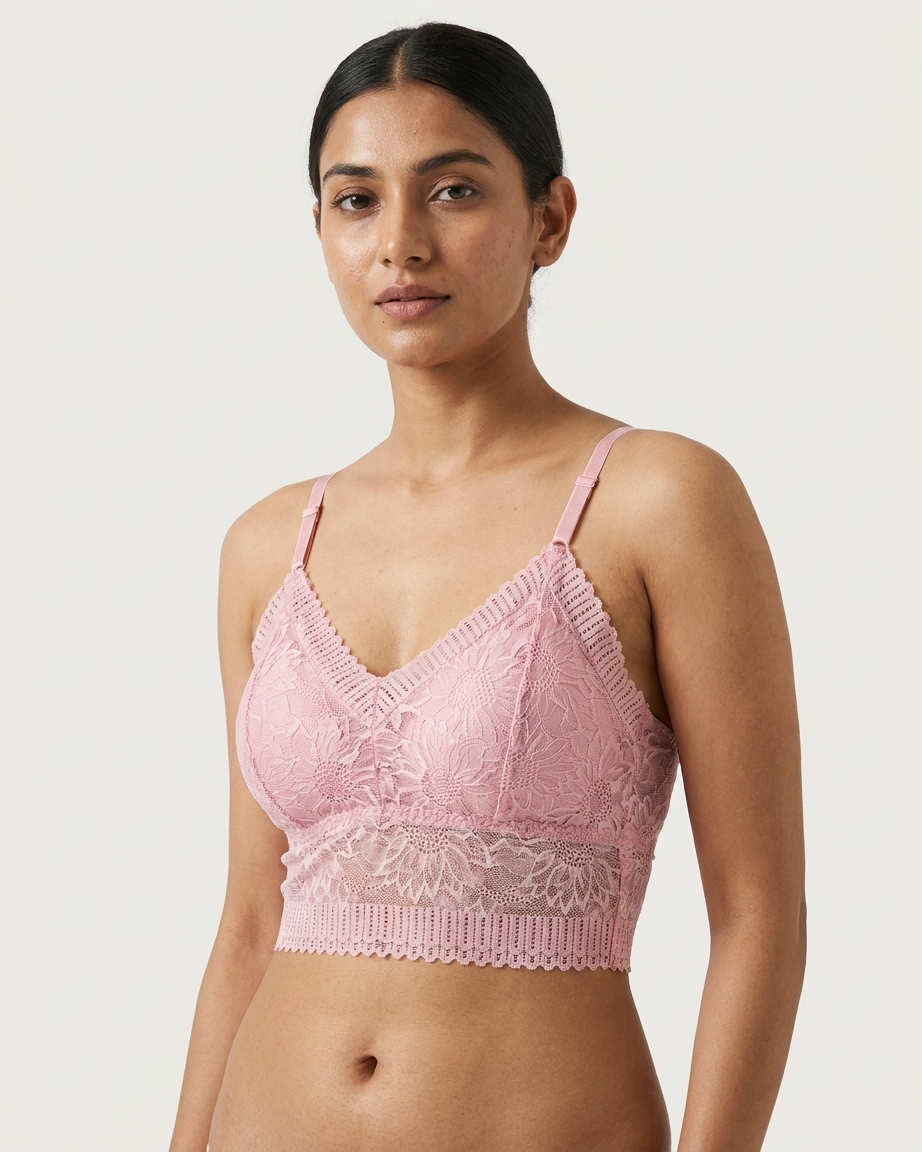 A woman modeling a light pink lace bralette with thin straps and a scalloped underband against a plain light gray studio background.