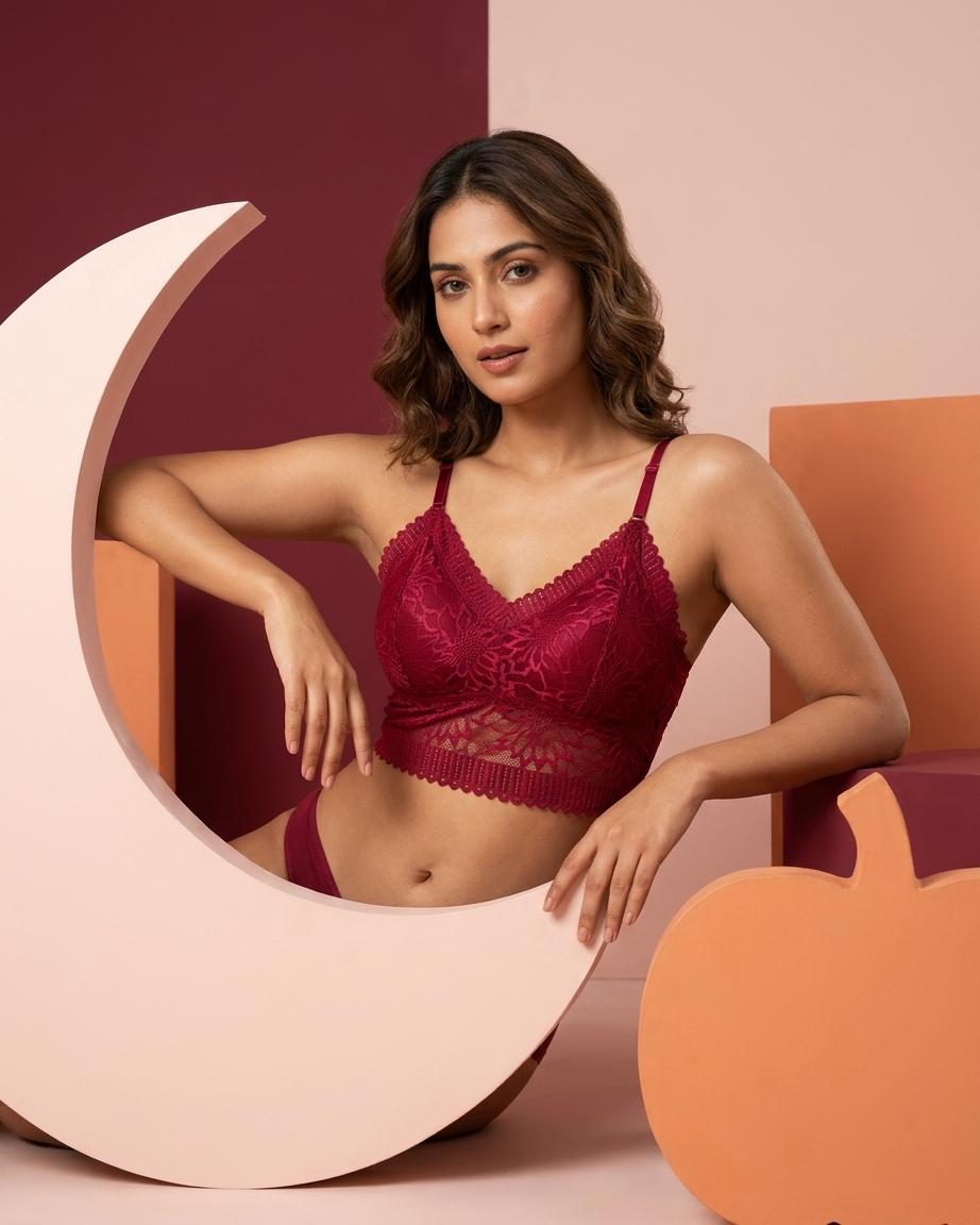 A woman models a deep burgundy lace bralette and matching underwear set, posed with large decorative props in a studio setting with a warm color palette.